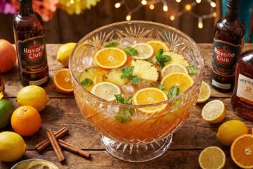 A large glass punch bowl filled with tropical punch, garnished with slices of orange, lemon, pineapple and fresh mint, surrounded by bottles of rum, whole citrus fruits and cinnamon sticks on a rustic wooden table.